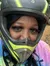 A person is taking a selfie while wearing a motocross helmet, with a tie-dye shirt and an ATV rider visible in the background.