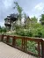The image captures a rustic and whimsical treehouse with bridges set amidst vibrant greenery, viewed from a wooden boardwalk.
