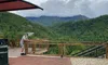 A person stands on a brick patio with a breathtaking view of green mountains under a cloudy sky.