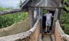 A person is standing in the doorway of a rustic wooden hut with a steeply pitched roof, accessed by a bridge with rope netting sides and log supports.