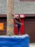 A person in a plaid shirt and jeans is engaged in a log climbing activity, using a rope and climbing spikes to ascend a tall pole, with a padded safety base below.