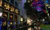 The image captures a vibrant evening scene at a riverside with a colorfully lit tree, architecture featuring balconies with glowing lights, and a dragon boat adding to the picturesque atmosphere.