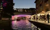 The image captures an evening scene of a vibrant urban riverwalk with pedestrians, market stalls, and an illuminated bridge reflecting colored lights onto the water.