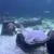 The image shows stingrays and fish swimming among underwater structures that resemble sunken debris in a clear aquatic environment, possibly an aquarium exhibit.