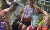 A happy family dressed in colorful outfits is enjoying a sunny day together, likely at an amusement park or public attraction, evidenced by the casual clothing, accessories, bench seating, and relaxed atmosphere around them.