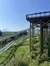 This image features a mountain coaster with tracks winding down a slope and an elevated platform set against a backdrop of lush greenery and distant mountains under a clear blue sky.