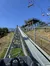 The image shows the view from a mountain coaster cart ascending a track with a ski lift running parallel under a clear blue sky.