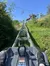 A person is riding a mountain coaster heading up an incline with an overhead chairlift and clear blue skies in the background.