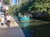 A boat full of passengers cruises along a river beside a walkway where a person is walking and kiosks display souvenirs.