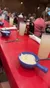 The image depicts a casual dining setting with a bowl of what appears to be clam chowder and a lemonade-like beverage on a red table, with blurred background activity indicating other diners.