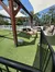 A miniature horse stands on artificial turf within an enclosure, beside a water trough and under a shade structure, with visitors observing from beyond the fence.