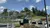 The image shows an outdoor scene with a mountain coaster track winding its way down a lush hillside, passing over a parking area with a few vehicles, under a clear blue sky with scattered clouds.