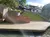 The image shows a sunny outdoor setting with a canal or river in the foreground, a paved embankment, and a grassy, stepped area leading up to white buildings with a sign saying