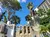 An urban scene featuring palm trees, a stone wall with decorative arches, a white building with balconies, and a high-rise in the background, under a clear blue sky.