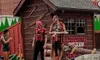 Three people are engaged in a lumberjack-themed performance or demonstration with props like logs and axes, set against a small wooden hut backdrop.