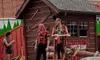 Three performers dressed in lumberjack-themed attire are interacting on a stage designed to resemble a rustic, outdoor setting with a log cabin and props like saws and logs.