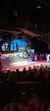 The image features an audience watching a live performance with performers on a stage set resembling a rustic outdoor scene, with tables in the foreground indicating a dinner theater setting.