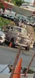 The image shows a vintage truck with a large sculpture of a cow on its bed, parked among other vehicles in a sunny outdoor setting.