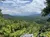The image captures a verdant mountainous landscape under a partly cloudy sky, with hints of human habitation tucked among the foliage and the corner of an off-road vehicle visible in the foreground.