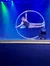 A performer is striking an athletic pose within a large ring against a blue stage backdrop with colorful stage lighting.