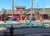 The image showcases an outdoor lumberjack-themed performance area with individuals engaged in axe throwing and sawing activities as a part of an entertainment show, with a small audience and bright blue skies overhead.
