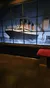 The image shows an exhibit display featuring a large photo of the Titanic traversing the ocean, with informational text on the right and a bench in the foreground for viewers.