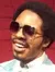 A person is seated at a piano smiling next to a wax figure of a man in a blue suit and sunglasses.