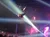 An aerial performer is executing a dramatic upside-down pose on a swing-like apparatus above an audience with colorful stage lighting enhancing the atmosphere.
