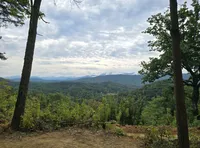 The image showcases a lush, rolling mountain landscape viewed from a forested area under a cloudy sky.