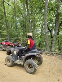 A person is sitting on an all-terrain vehicle (ATV) on a dirt path in a forested area.