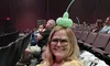 A smiling person is seated in a theater wearing glasses and a balloon hat while holding yellow balloon sticks with audience members and theater seating in the background.