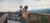 A person is holding a child while both gaze out over a scenic mountain view from a high vantage point.