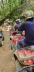 A line of people on ATVs wearing helmets are trailing through a forest trail.