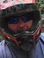 Two people wearing helmets are on an outdoor adventure, with one taking a selfie and the other sitting on an ATV in a forested area.