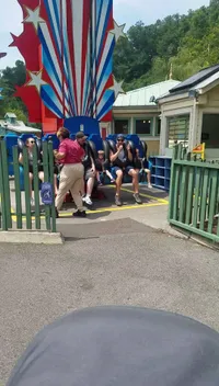 A group of people is seated on a star-spangled amusement park ride, with some participants looking excited and an attendant helping to secure a rider.