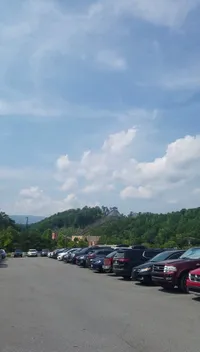 A parking lot is full of cars with a backdrop of a lush green hill and a clear, partially cloudy sky.