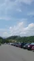 A parking lot is full of cars with a backdrop of a lush green hill and a clear, partially cloudy sky.