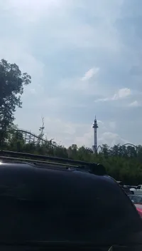 The image shows a sunny day with a clear blue sky, a view of a roller coaster track, and the tip of a drop tower ride, seen from a parking area with several cars.