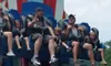A group of people, including adults and children, are seated on a drop tower amusement park ride against a backdrop with bright red, white, and blue patterns.