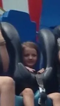 A group of people, including adults and children, are seated on a drop tower amusement park ride against a backdrop with bright red, white, and blue patterns.