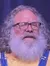 A man with a white beard, wearing glasses, a yellow shirt, and blue overalls is standing on a stage with a puppet resting on a stool beside him, in front of a closed curtain with blue and yellow lights.