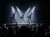 The image shows a live drumming performance on a stage with dramatic backlit lighting, captivating an audience sitting in the darkened theater.