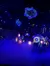 The image shows a colorful theatrical performance with large lit-up flower props and floating fabric elements, creating a whimsical, dream-like atmosphere, likely observed by a live audience.
