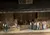 The image shows a group of actors on stage dressed in historical costumes, suggesting a scene from a period play or a historical reenactment, set against a rustic, barn-like backdrop with props like hay bales and a table with a gingham cloth.