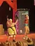 Two actors on stage appear to be engaged in a comedic skit, with one brandishing a pan as a mock weapon and the other reacting in exaggerated fear.