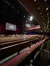 The image shows the interior of the Grand Ole Opry with rows of empty seats and a few scattered audience members, bathed in warm ambient lighting with a stage in the background displaying the Opry logo.