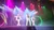 A group of performers in white and black attire is singing and dancing on a stage with colorful lights and hazy effects.