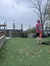 A person in shorts and a cap stands on an artificial grass lawn scattered with fallen leaves, looking toward the horizon, while another person stands in the background near outdoor furniture.