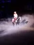 A person wearing a traditional rodeo outfit rides a horse through a dimly lit arena with a dramatic shadow cast behind them.