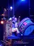 A performer in an ornate costume stands next to a large, colorful drum on a dimly lit stage with stage lighting overhead.